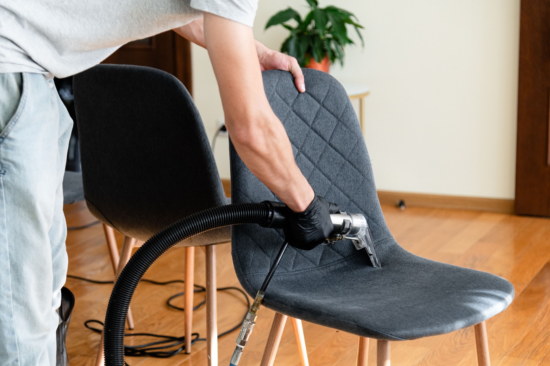 A person cleans a dark upholstered chair using a specialized cleaning tool in a well-lit room. The wooden floor and minimalist decor